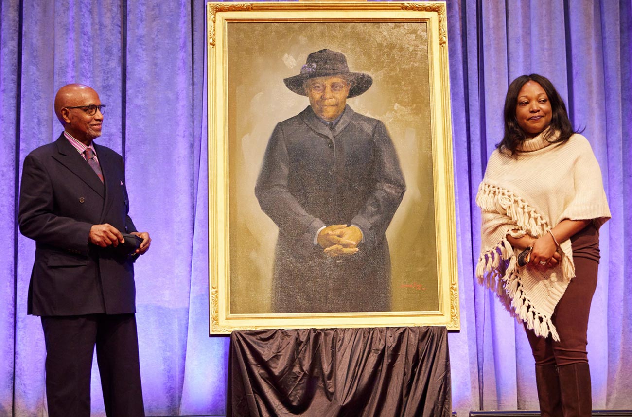 a man and woman stand on a stage on either side of a painted portrait of Lucille "Lucy" Byard