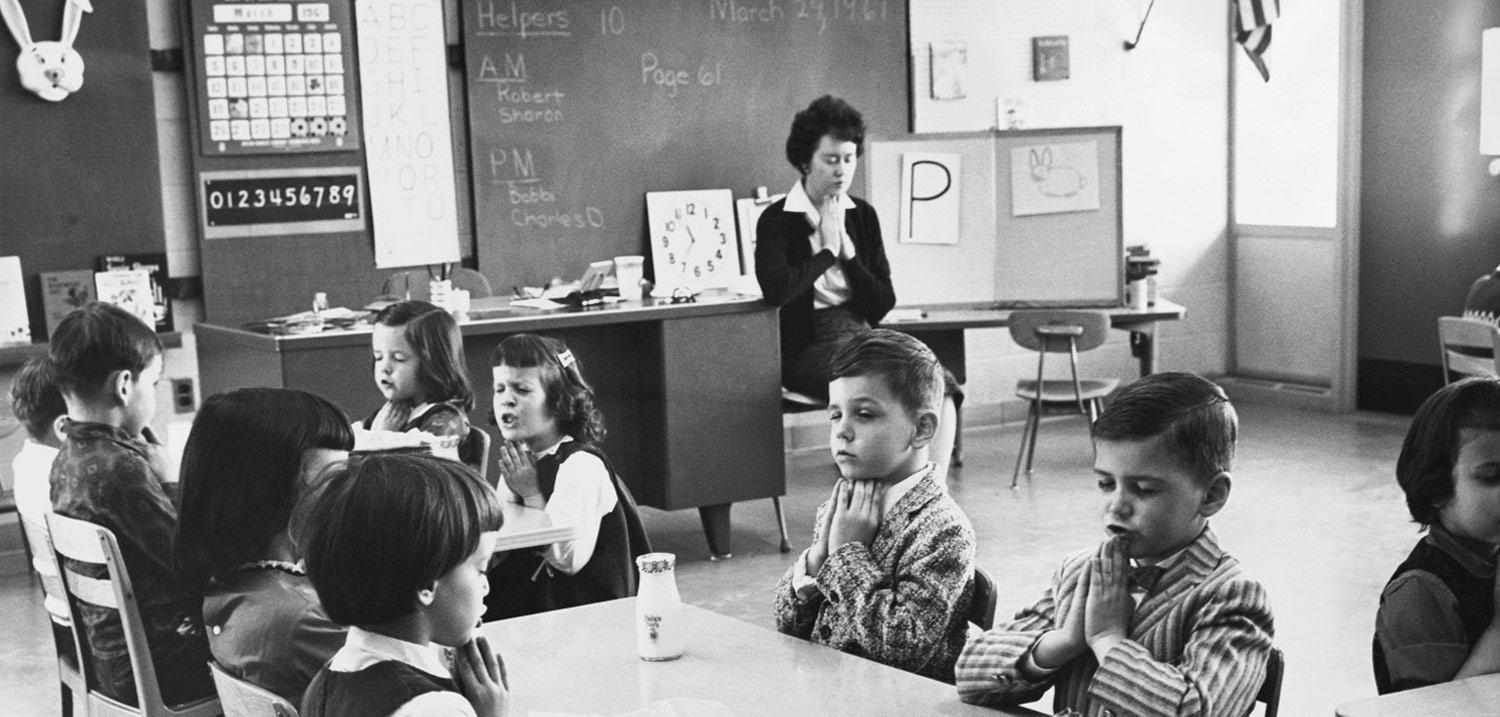 an old black and white image of a teacher leading her young class in prayer