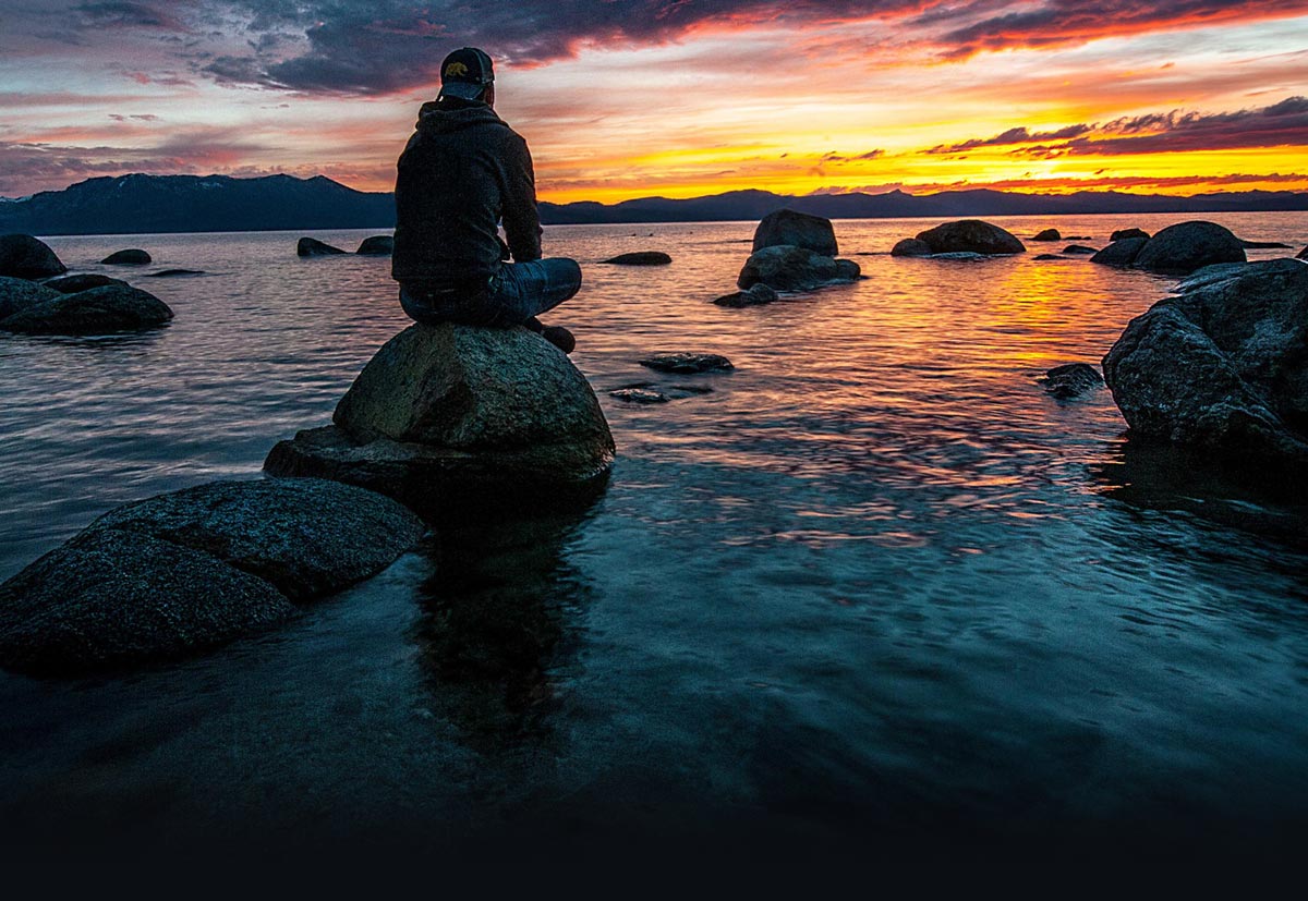 a man watches the sunset while sitting on a boulder on the beach