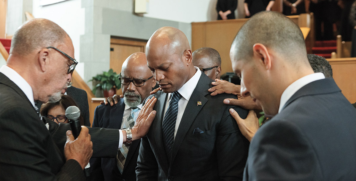 a group of people stand around Governor Wes Moore, resting their hand on his shoulders with their heads bowed in prayer