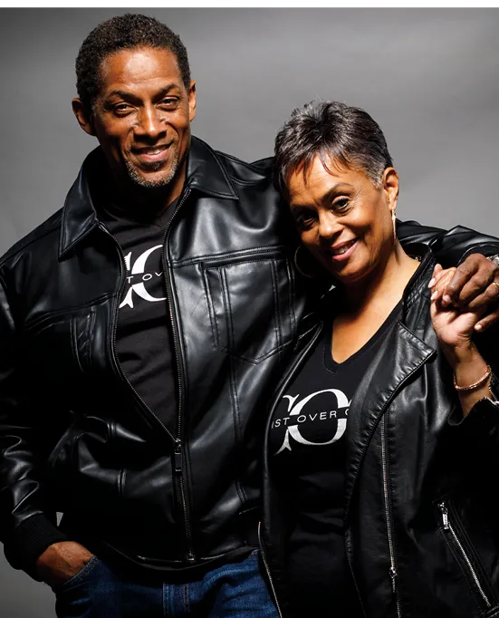 A smiling man and woman in matching black leather jackets, standing together. Both are wearing black t-shirts with a circular logo on the front. The woman is holding the man's arm.