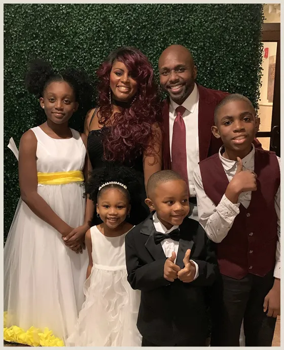 A family portrait featuring two adults and four children, all dressed in formal attire. The man is wearing a red suit jacket, and the woman is in a black dress. The children are dressed in white and black formal wear, with the youngest child smiling at the front.