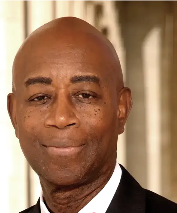 A close-up portrait of a bald man in a suit, smiling softly. The background shows blurred columns of a building.