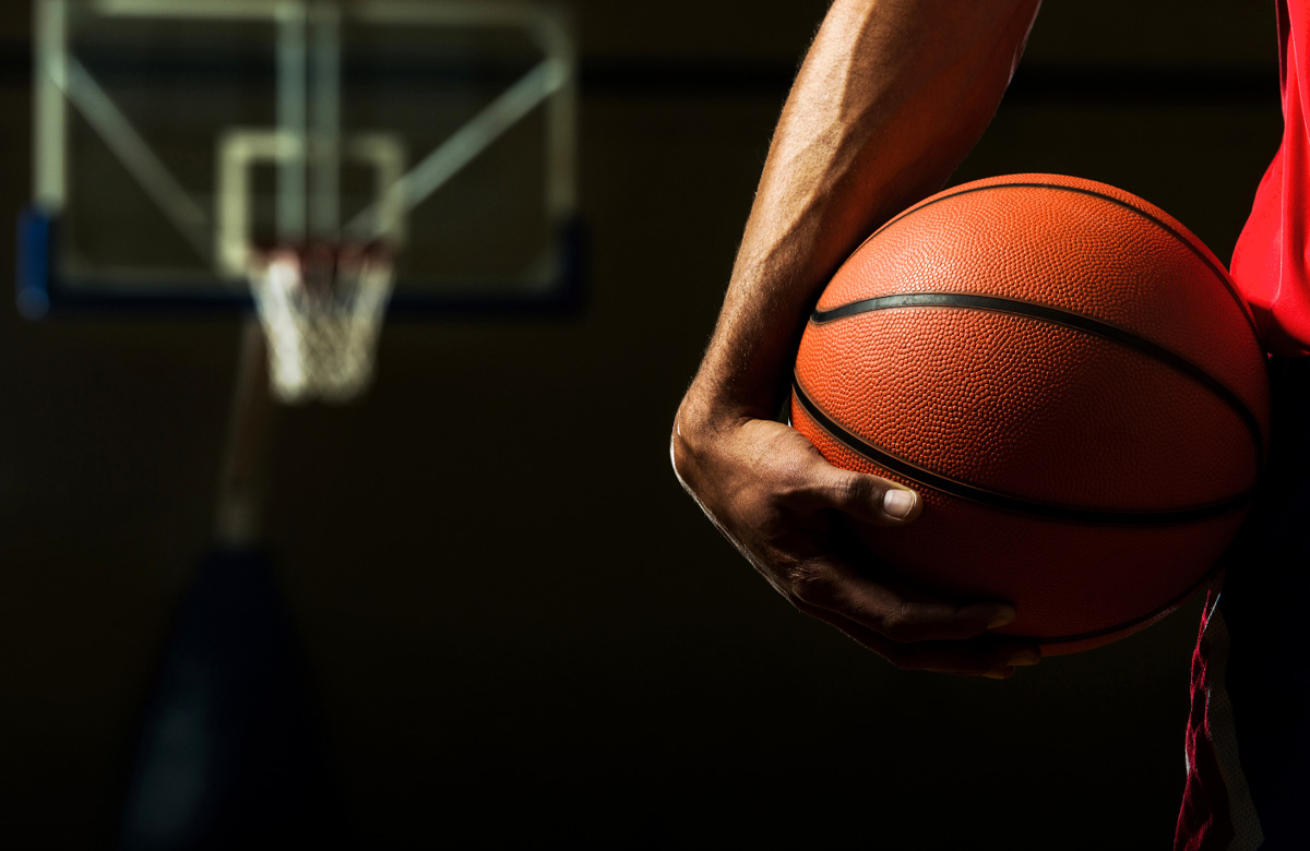 low cropped view of a hand holding a basketball with a hoop out of focus in the background