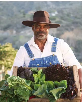Smiling Black man wearing overalls and a wide-brimmed hat holding a large wooden crate filled with freshly harvested leafy greens, standing outdoors with a scenic backdrop.