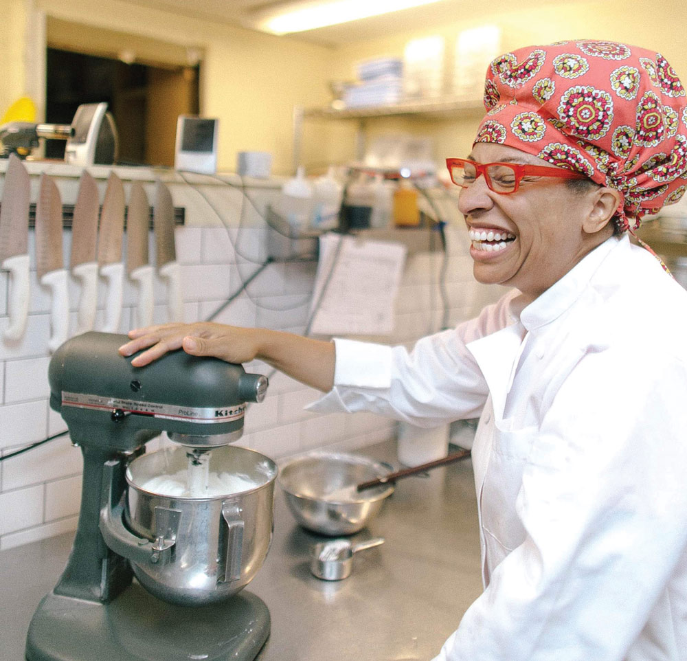 A smiling person in a chef's jacket using a stand mixer in a kitchen.