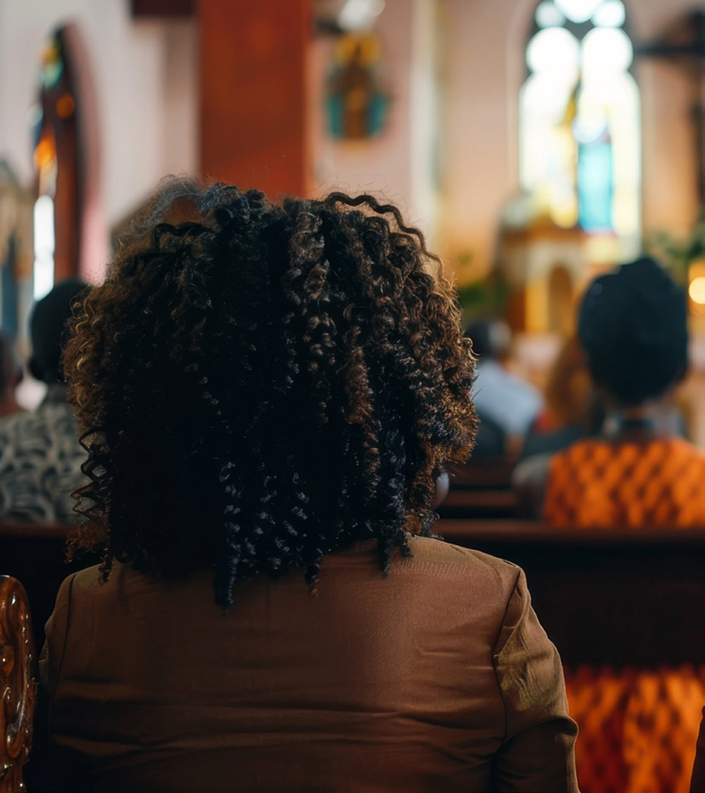 woman with curly brown hair sits in a church pew, viewed from behind