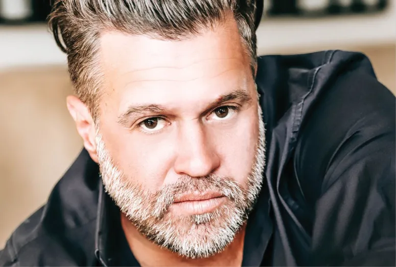 Close-up portrait of a man with salt-and-pepper hair and beard, wearing a navy blue shirt. He is gazing directly at the camera with a serious expression. The background is softly blurred.