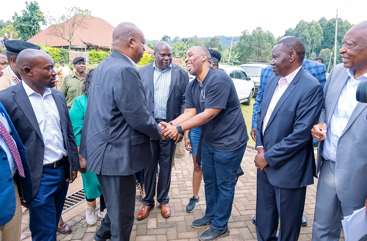 Two men in the center of a diverse group shake hands and smile, while other individuals in suits and casual wear observe outdoors.