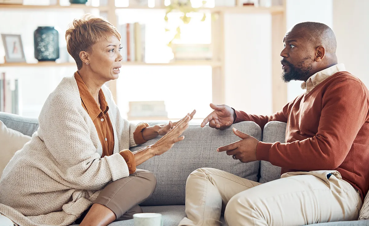 A Black couple sits on a sofa, engaged in a serious conversation, with the woman gesturing as she speaks to the man.