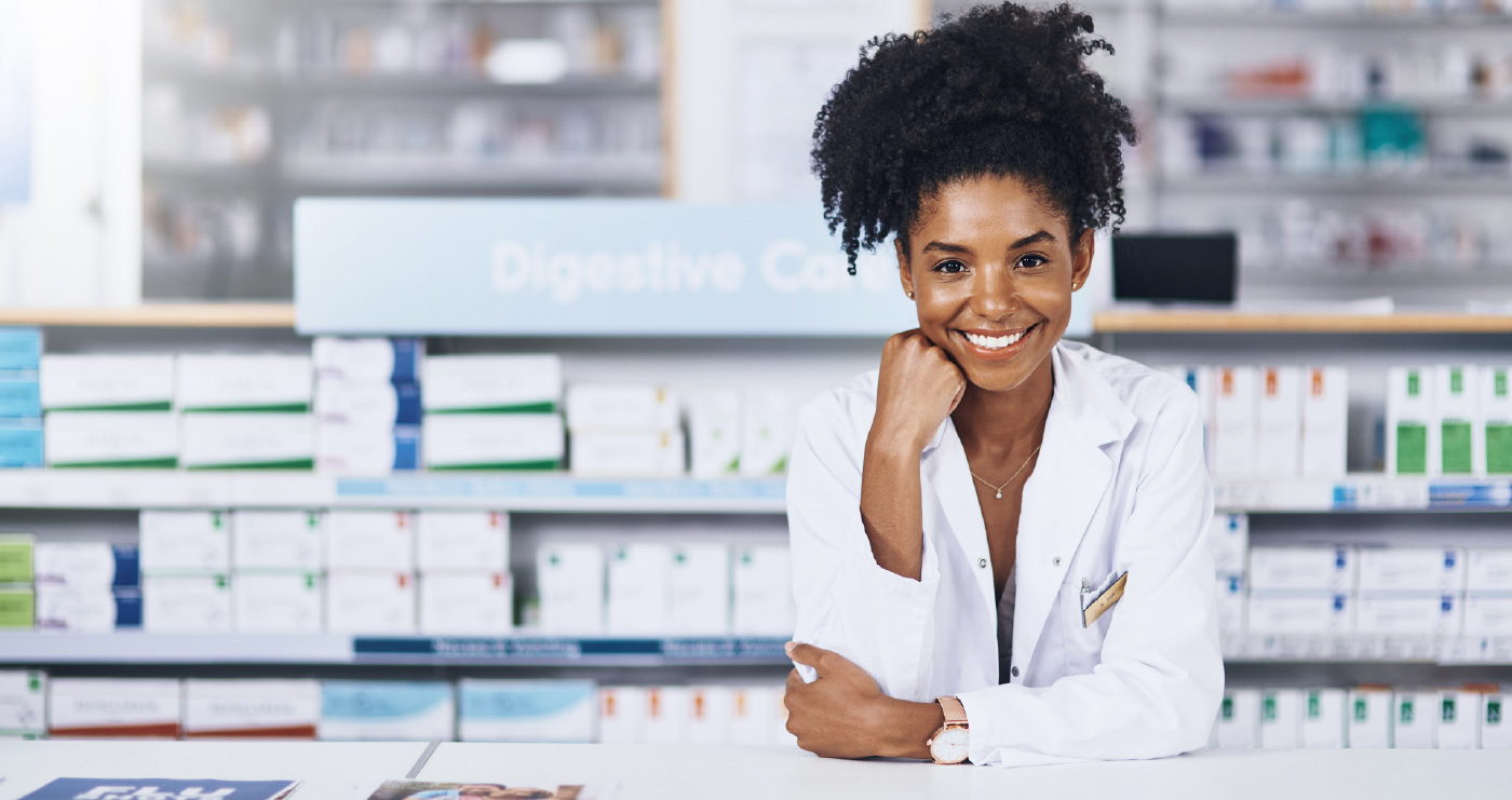 A friendly female pharmacist wearing a white lab coat, leans on a counter in a pharmacy while smiling
