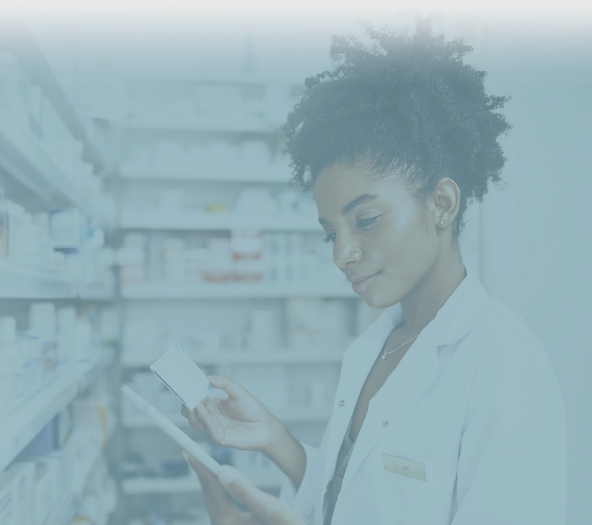 A female pharmacist wearing a white lab coat, is in a pharmacy aisle. She is holding a small white box in one hand and a digital tablet in the other, appearing to compare information.