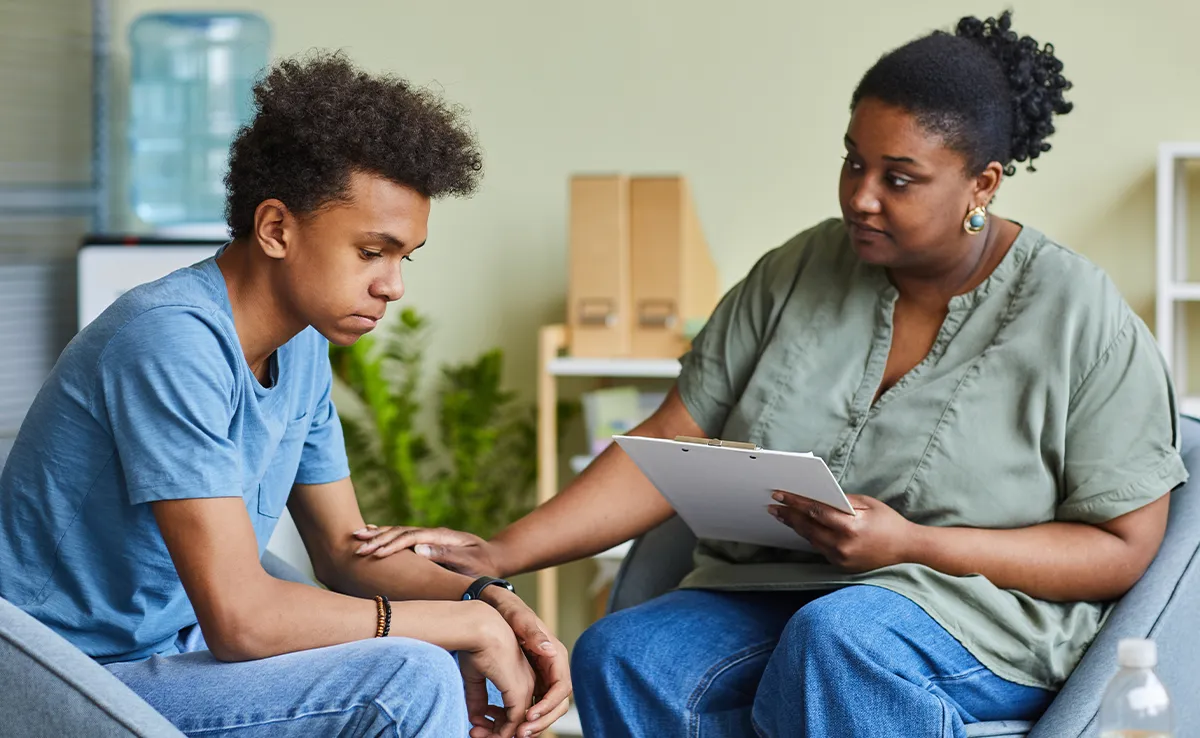 A Black female therapist or counselor sits across from a sad, teenage boy in a professional office setting. She is holding a clipboard and has her hand on his arm in a comforting gesture. The teenager, with his head down, appears to be listening intently.