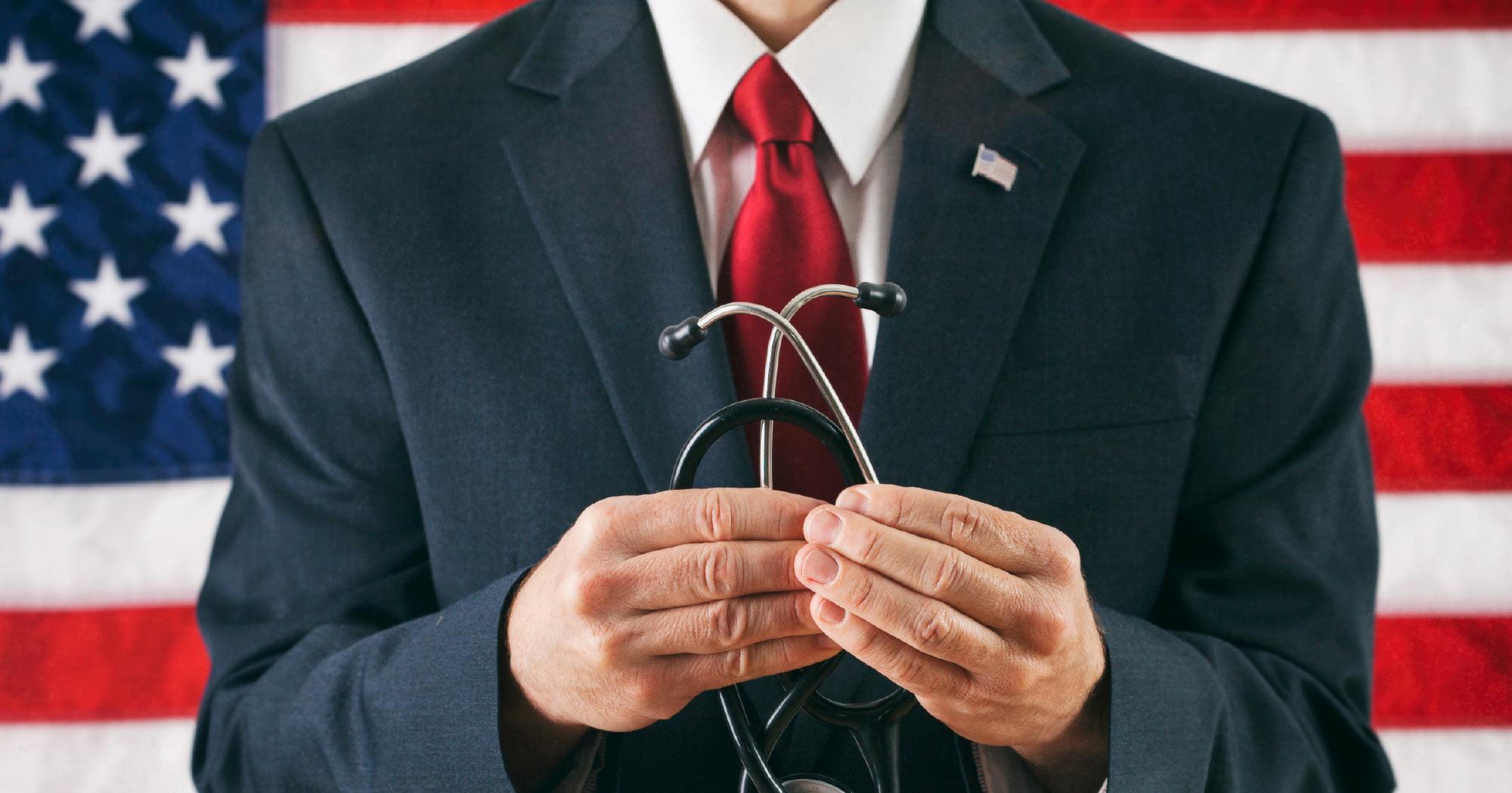 An eye-level, medium shot shows an adult male in a dark suit, white shirt, and red tie holding a black stethoscope with both hands in front of his chest. He wears an American flag pin on his lapel, and a large American flag serves as the backdrop. The man's head is not visible.