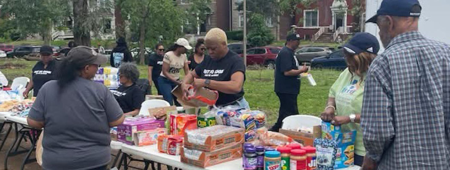 A group of people are gathered around a table with a variety of food items. The scene appears to be an outdoor community event, possibly a food drive or distribution. The background shows some trees and houses.