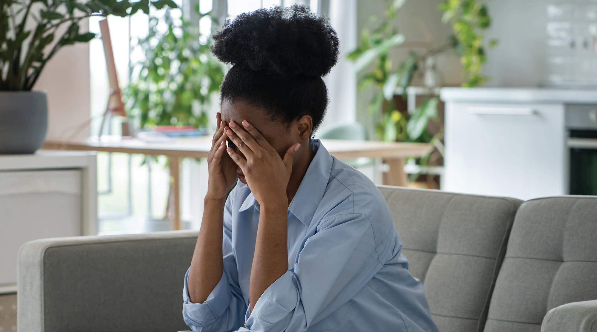 A photo of a woman sitting on a light gray sofa in a brightly lit, modern room. She is wearing a light blue shirt and has her hair pulled up in a bun. Her face is obscured as she covers it completely with both hands, conveying feelings of distress, frustration, or sadness.