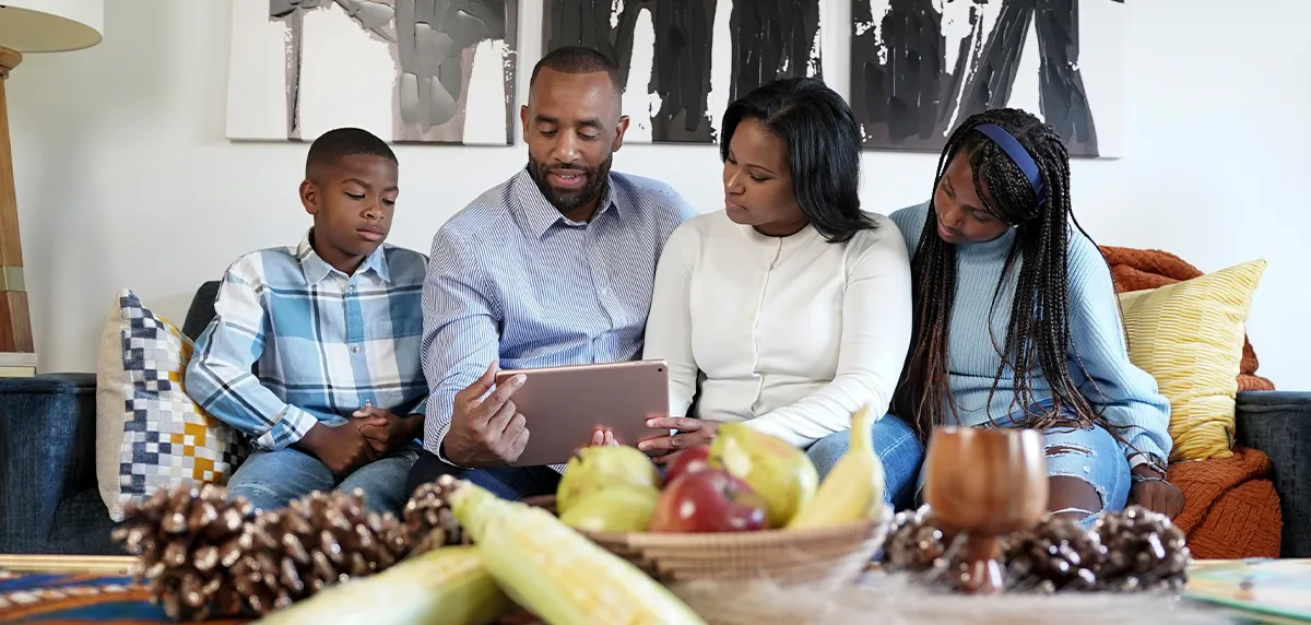 A family of four sitting together on a couch, looking at a digital tablet while relaxing in a modern living room filled with organic decor.