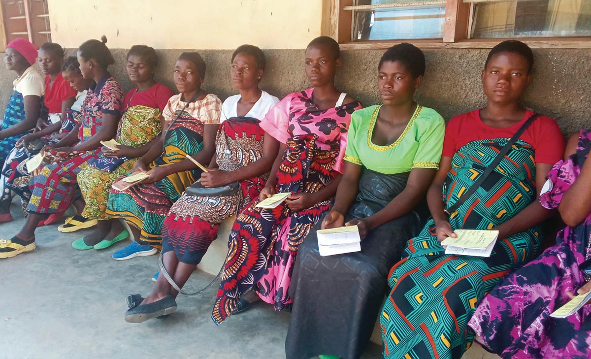 A row of women sit on a bench outside a clinic in Malawi, holding yellow health passports. They wear colorful traditional patterned wraps and shirts while waiting for medical services.