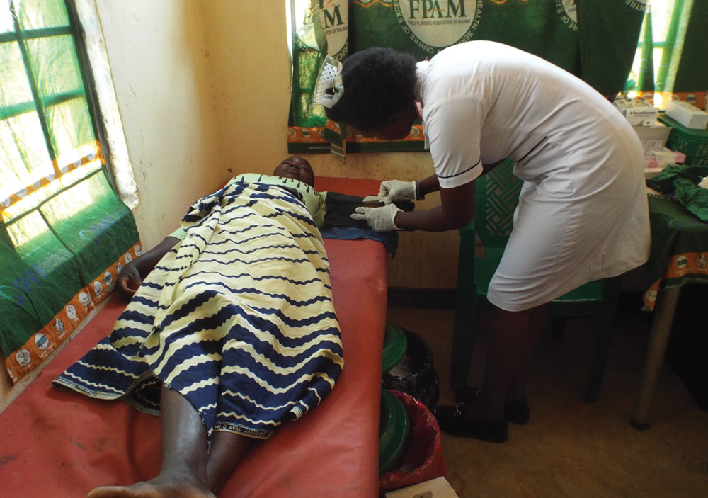 A nurse in a white uniform and gloves attends to a patient lying on a clinic bed. The room features green FPAM (Family Planning Association of Malawi) banners used as window coverings.