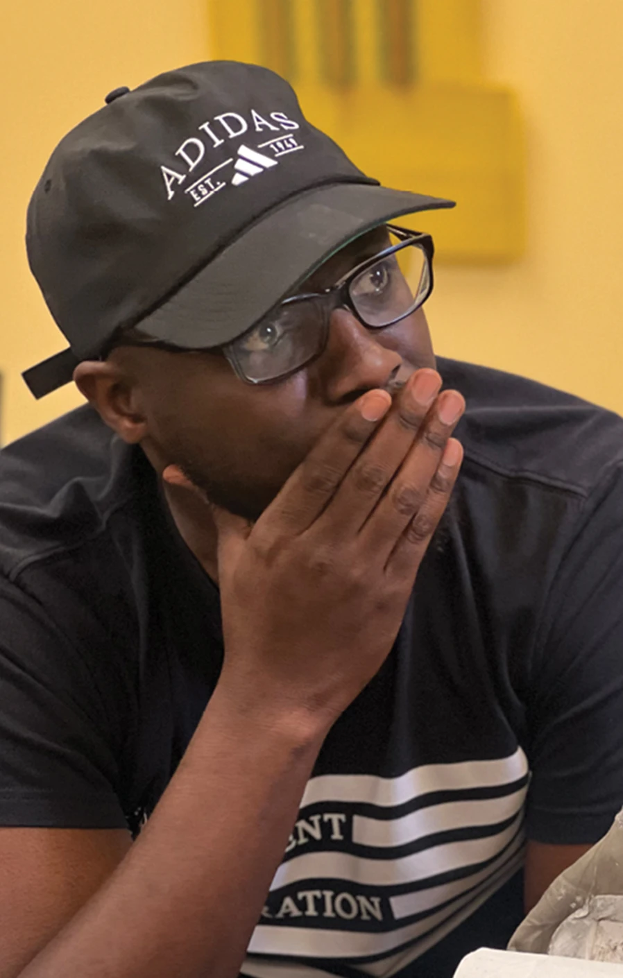 Man in glasses and an Adidas cap listening with his hand near his mouth.