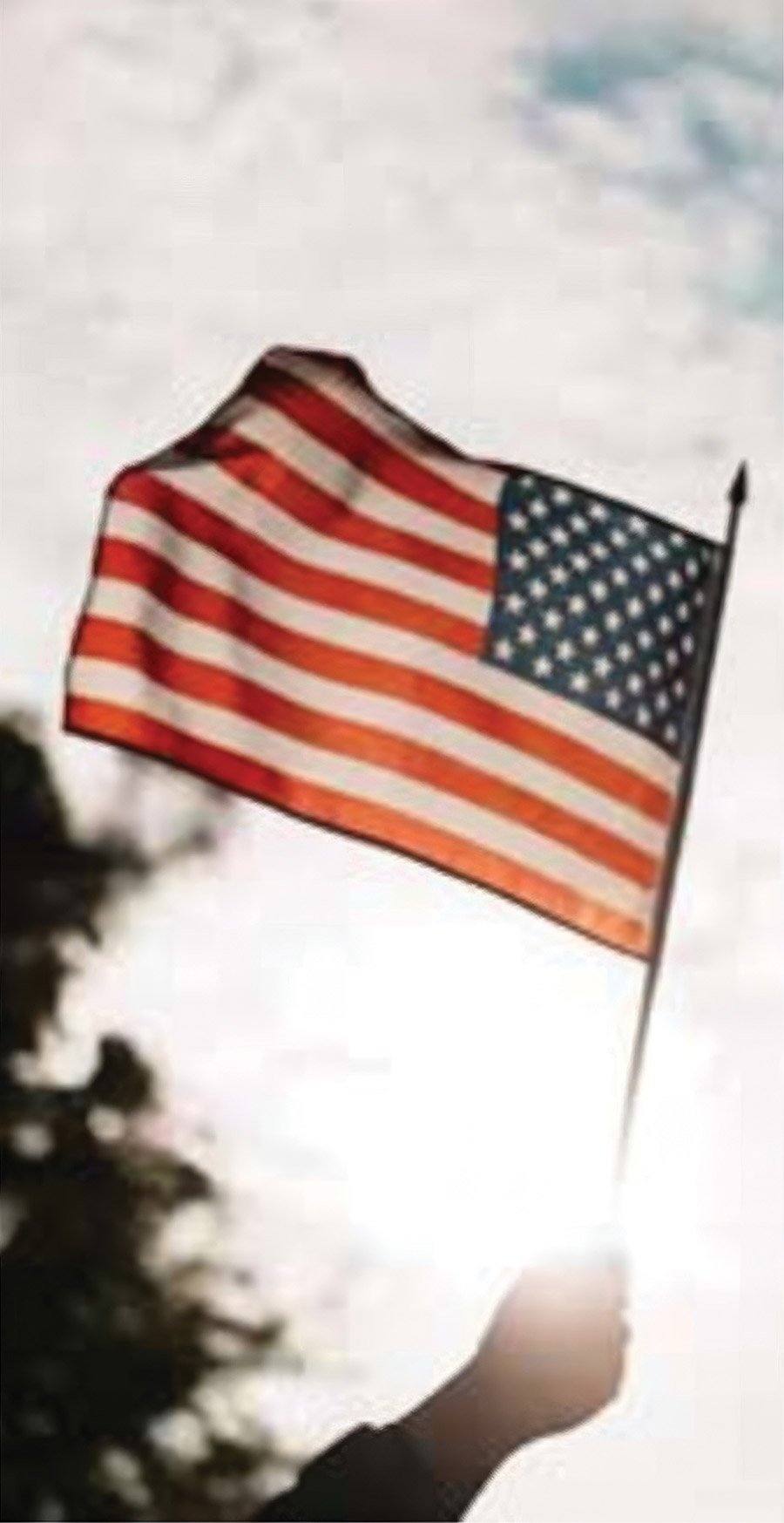 A hand holding up an American flag waving against a cloudy sky.