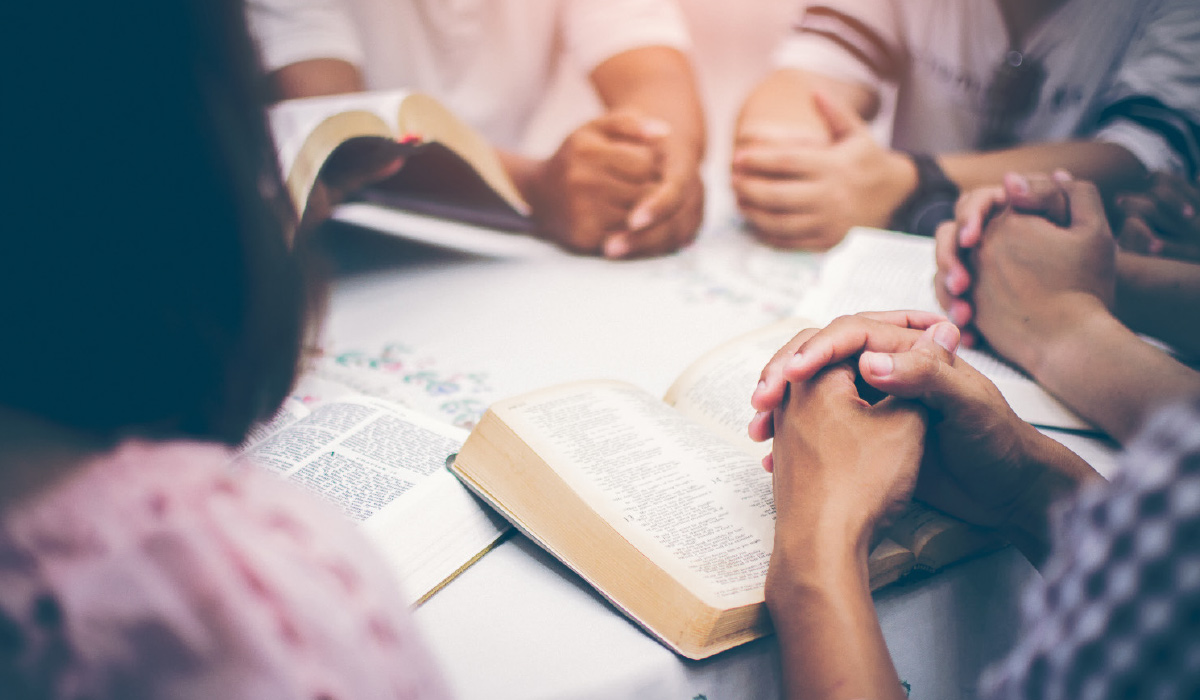 A close-up view of hands clasped together in prayer over open Bibles during a small group study around a table.