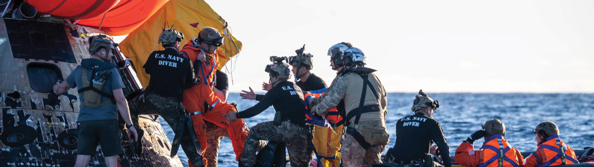 U.S. Navy divers assist individuals in orange suits near a floating spacecraft in the ocean.