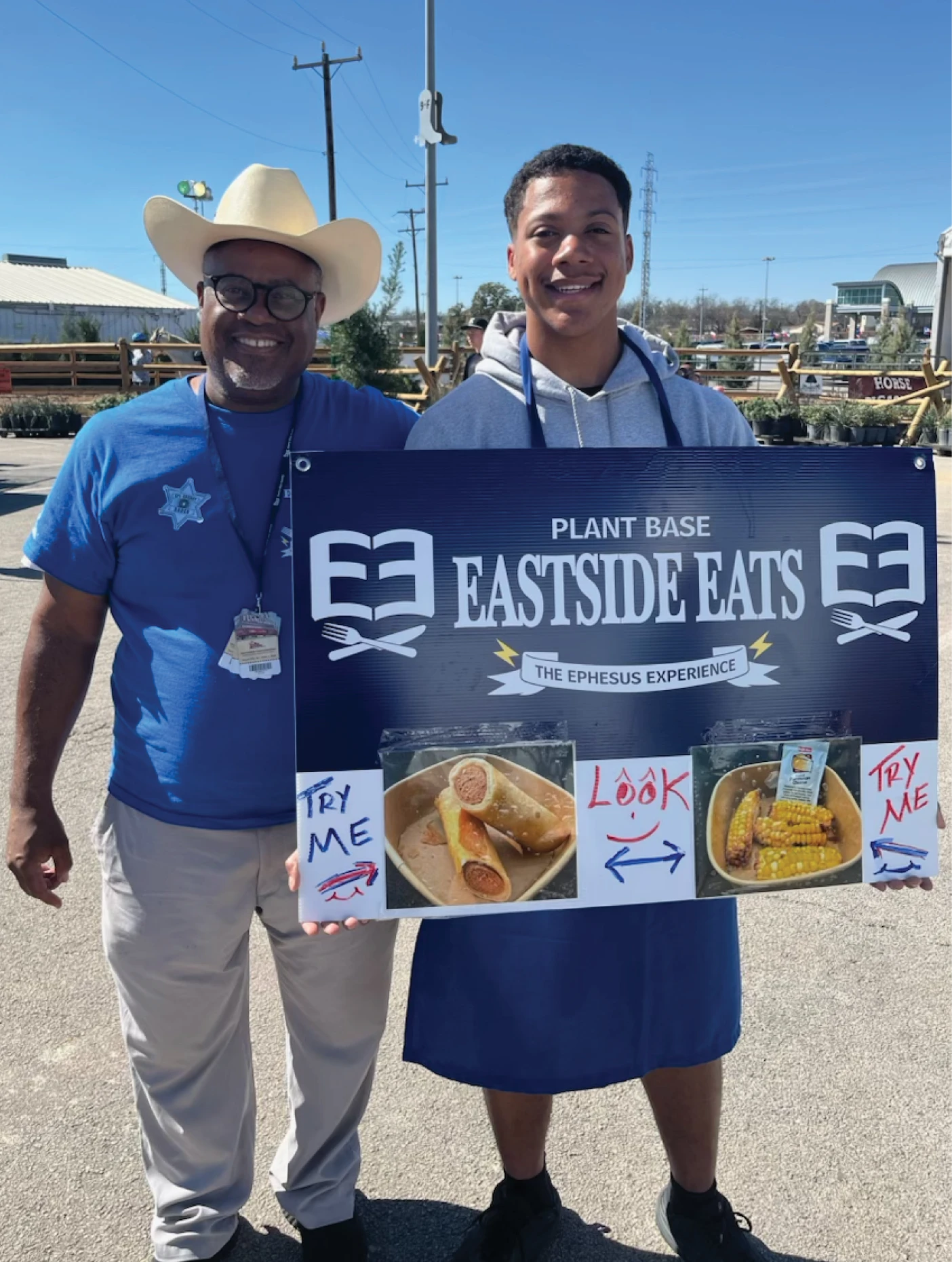 Two Eastside Eats volunteers holding a plant-based food sign at an outdoor event.
