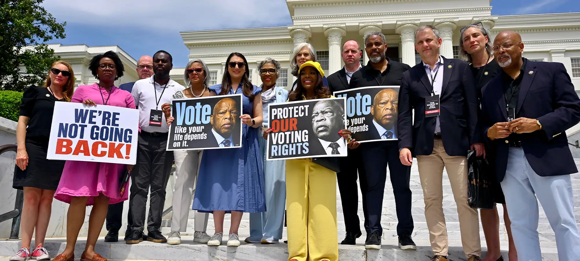 A diverse group of people stands on the steps of a white neoclassical building holding signs that read "We're Not Going Back!" and "Protect Our Voting Rights," featuring portraits of civil rights leader John Lewis.