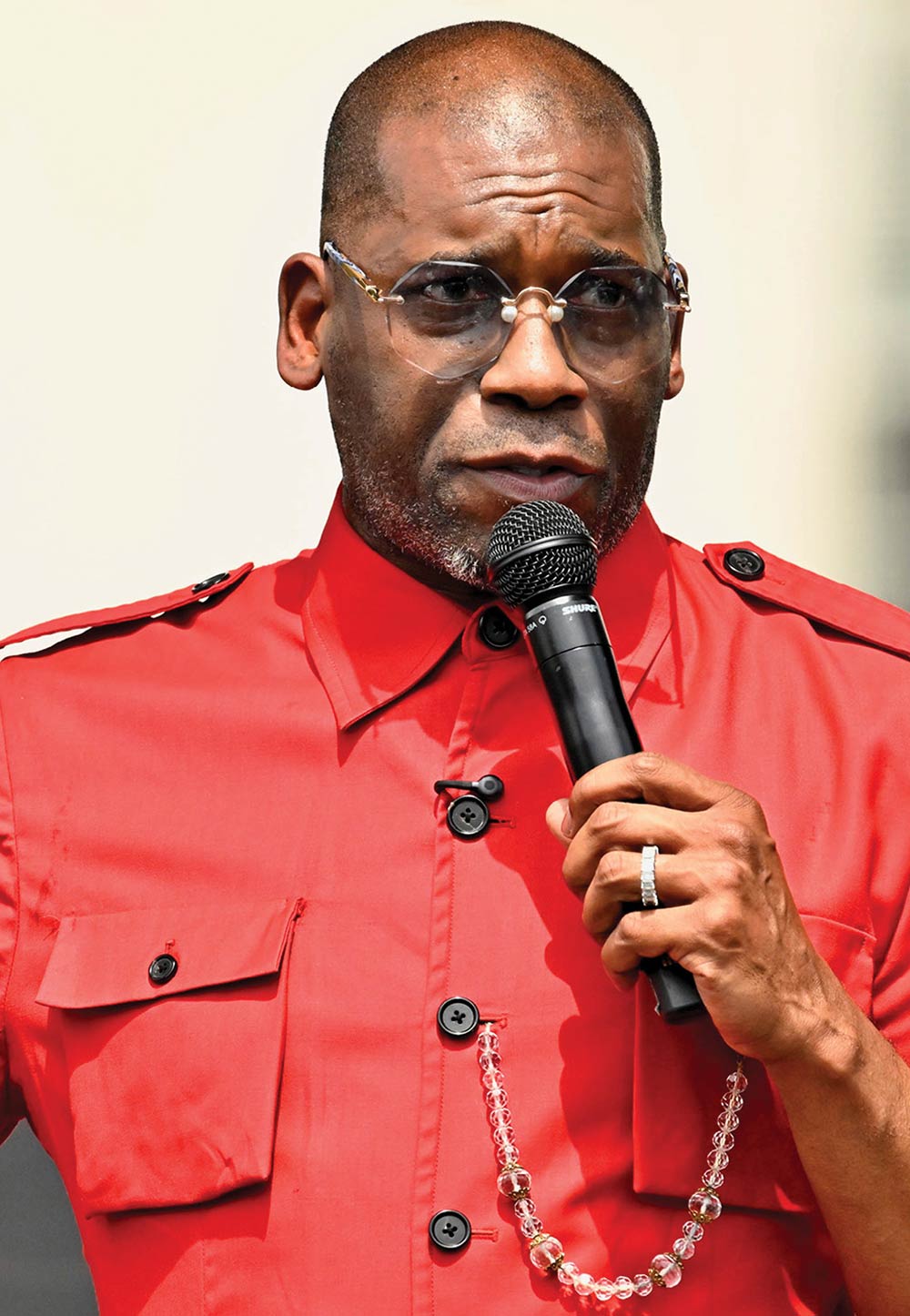 A Black man with rimless glasses and a short beard speaks into a handheld microphone while wearing a bright red button-down shirt with shoulder epaulets and a decorative beaded chain.