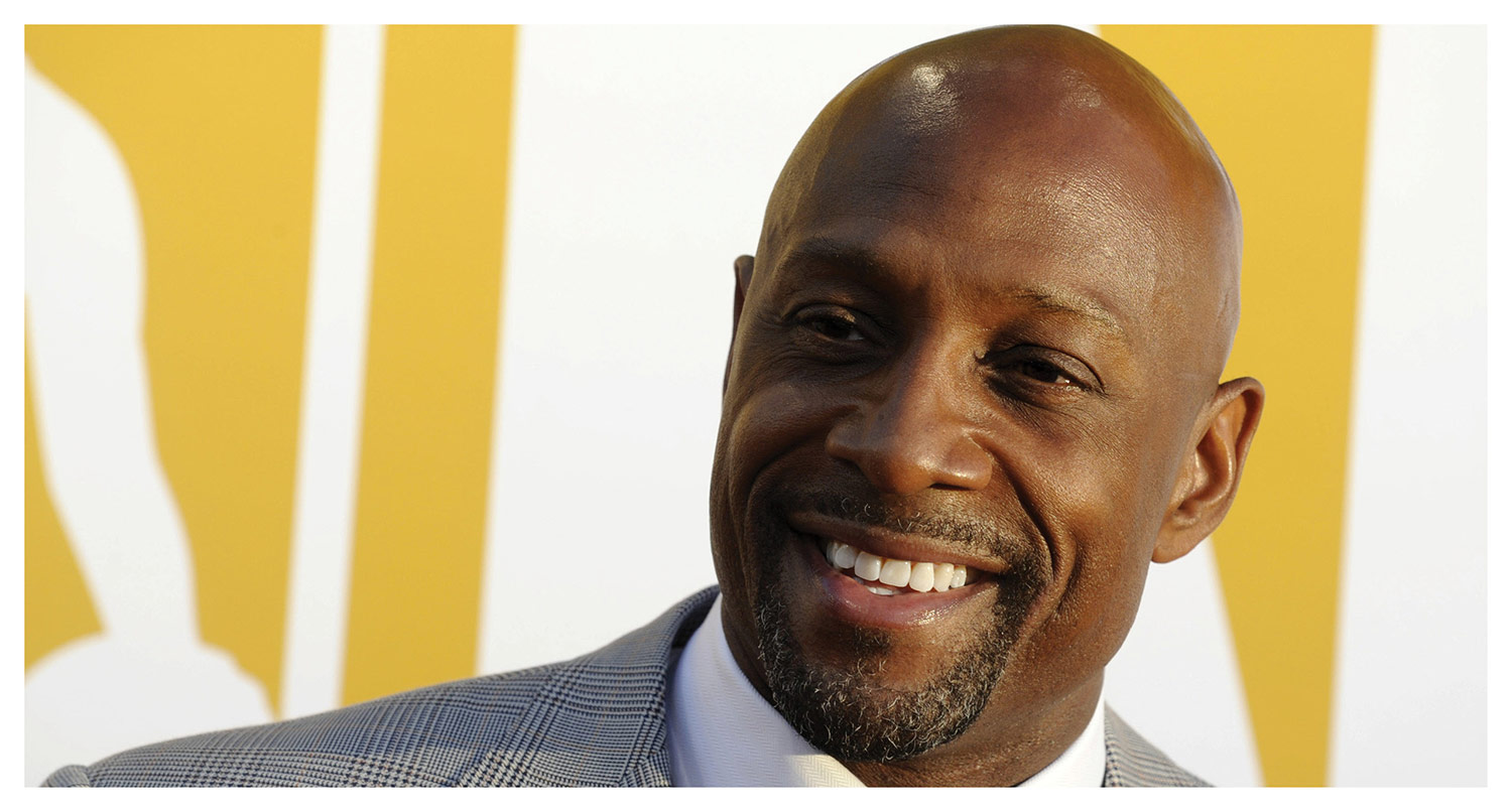 A close-up portrait of a smiling Black man with a shaved head and goatee, wearing a grey plaid suit jacket and white collared shirt against a yellow and white background.