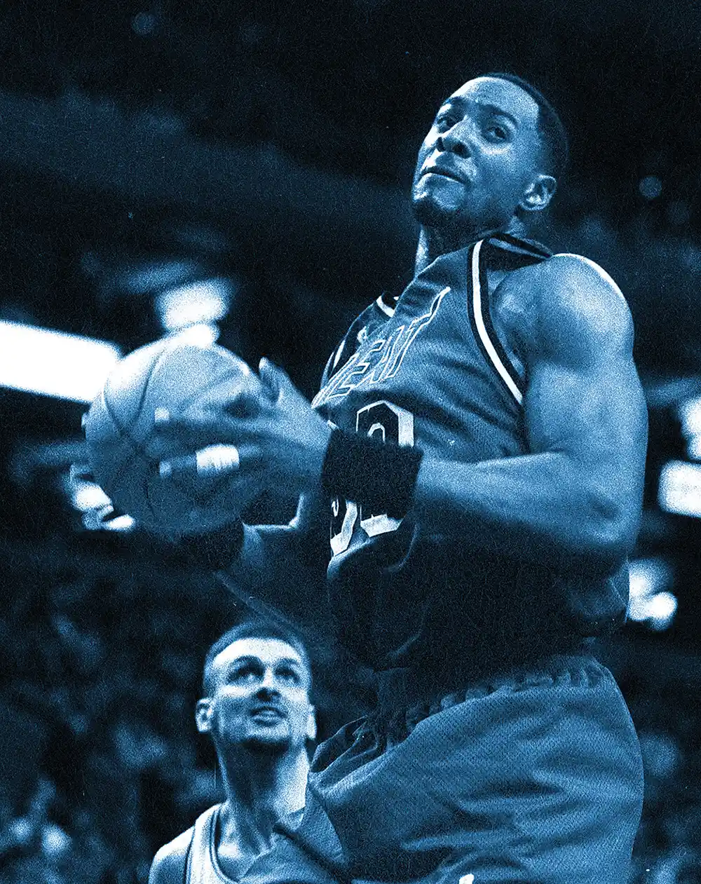 A blue-toned duotone photo of a professional basketball player holding the ball while mid-air during a game, with another player looking up from below.