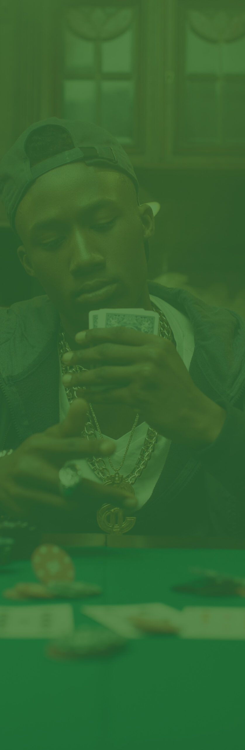 Green-tinted photo of a man wearing gold chains, holding a hand of cards at a poker table with poker chips.