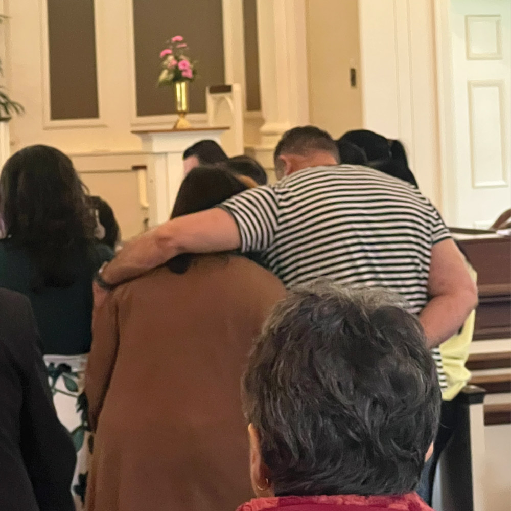 Man in a striped shirt comforting a woman with his arm around her shoulder inside a church.