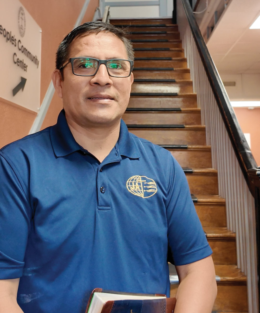 Man wearing glasses and a blue polo shirt standing in front of a wooden staircase while holding a book.