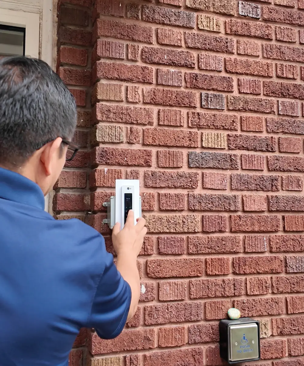 Man in a blue shirt using an electronic keypad mounted on a brick wall above an accessible door opener.