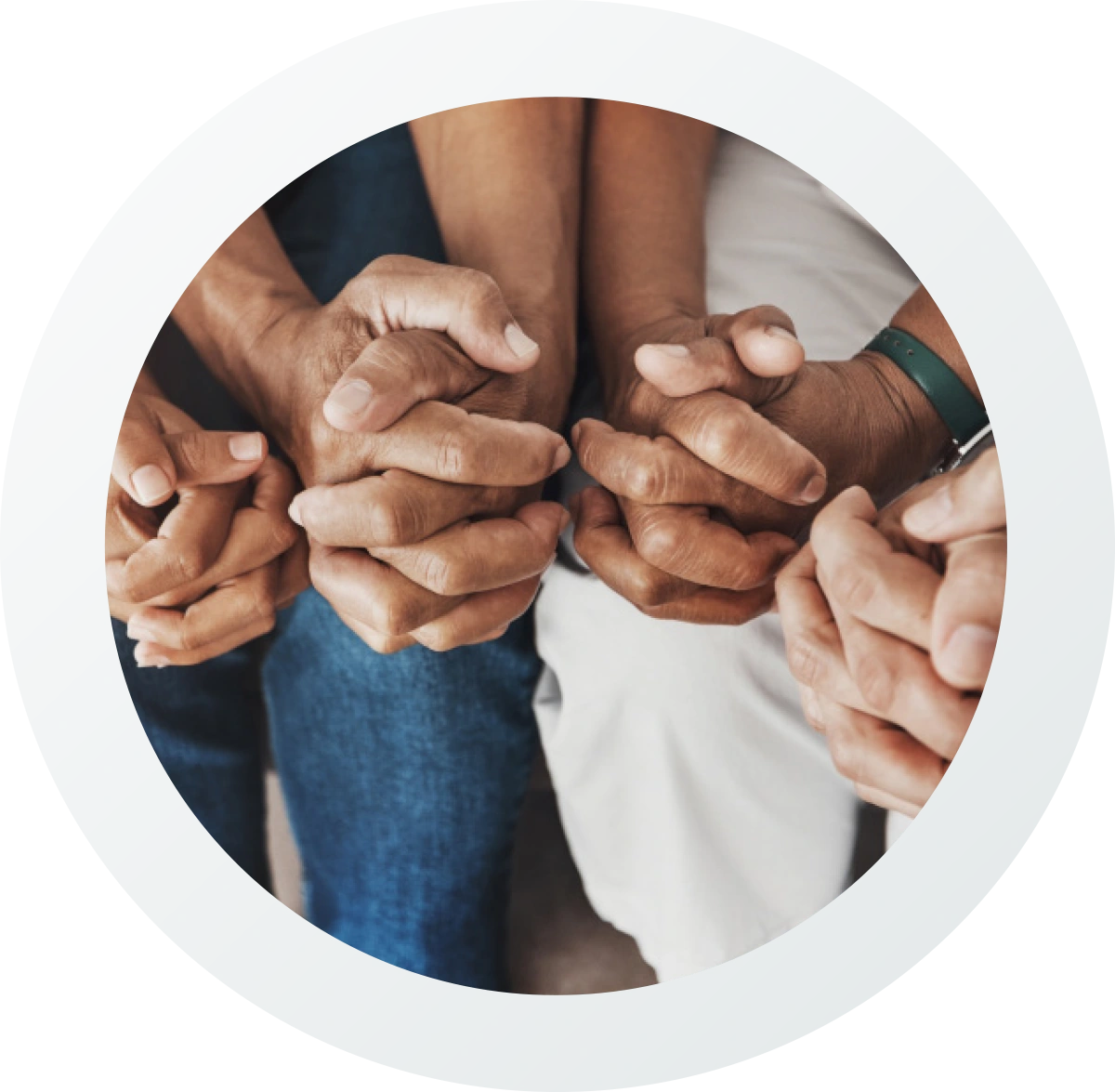 A circular photo of two people sitting, their clasped hands tightly locked and resting in their laps. One wears blue jeans, the other wears light-colored pants and a green bracelet. The pose represents unity, support, or prayer.
