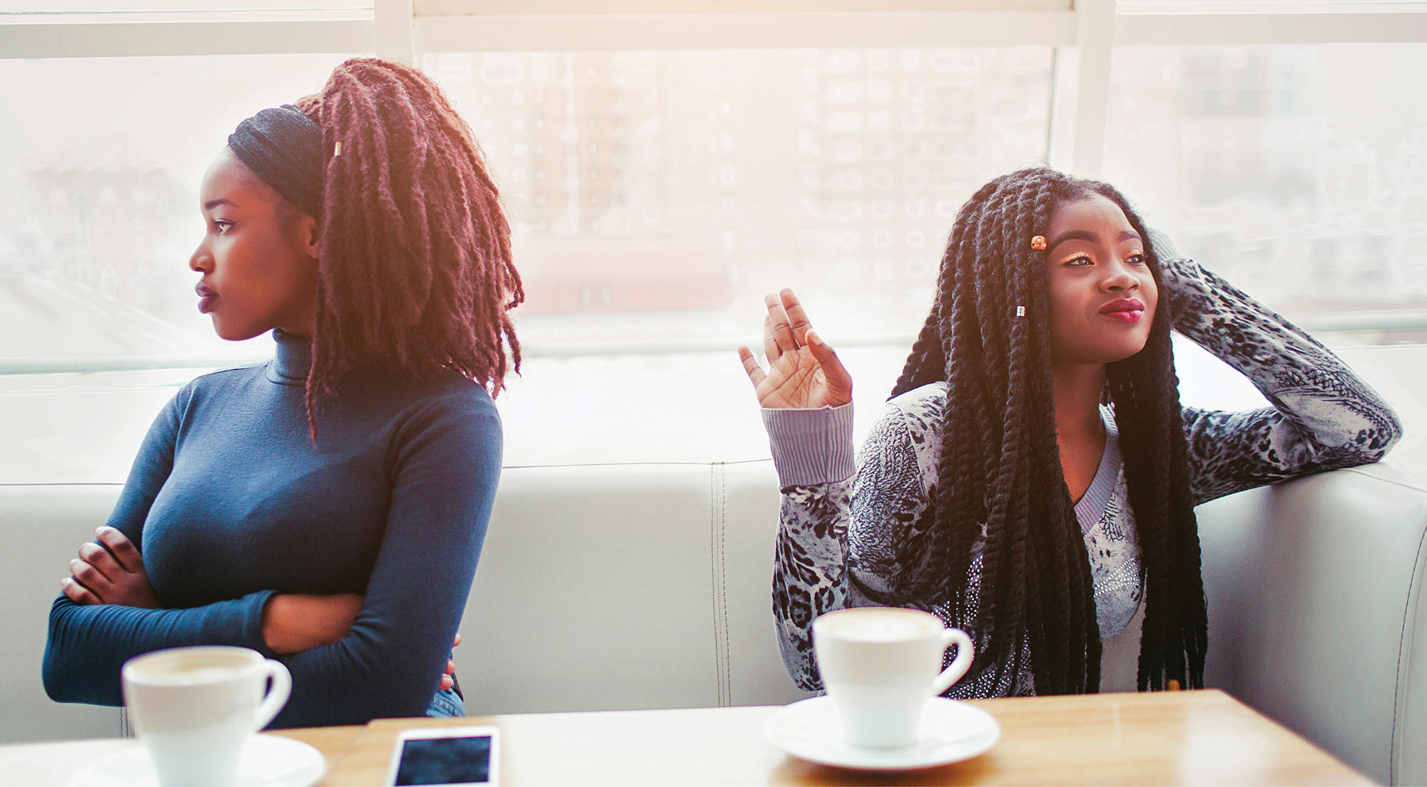 Two women sitting at a restaurant booth with coffee cups, turned away from each other during a disagreement.