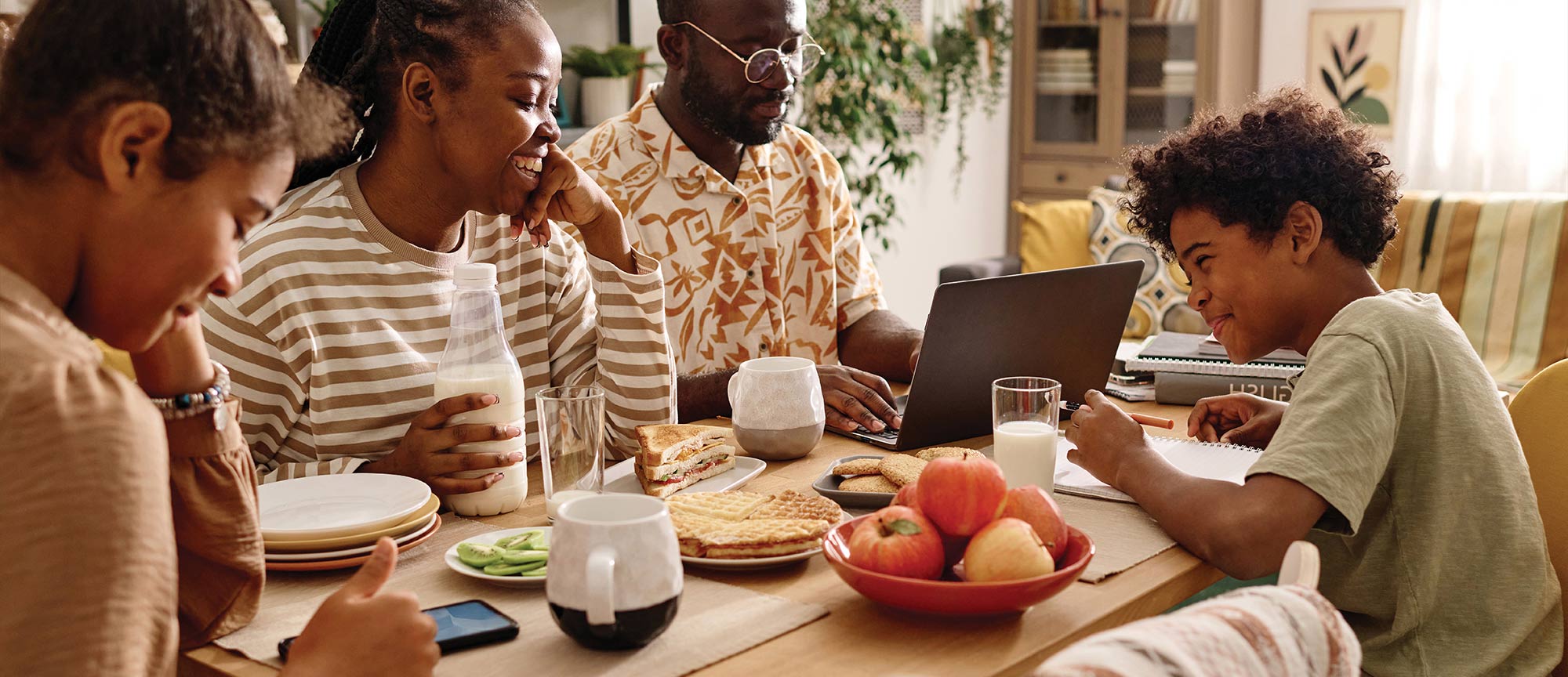 A family sitting around a table with food, milk, and a laptop.
